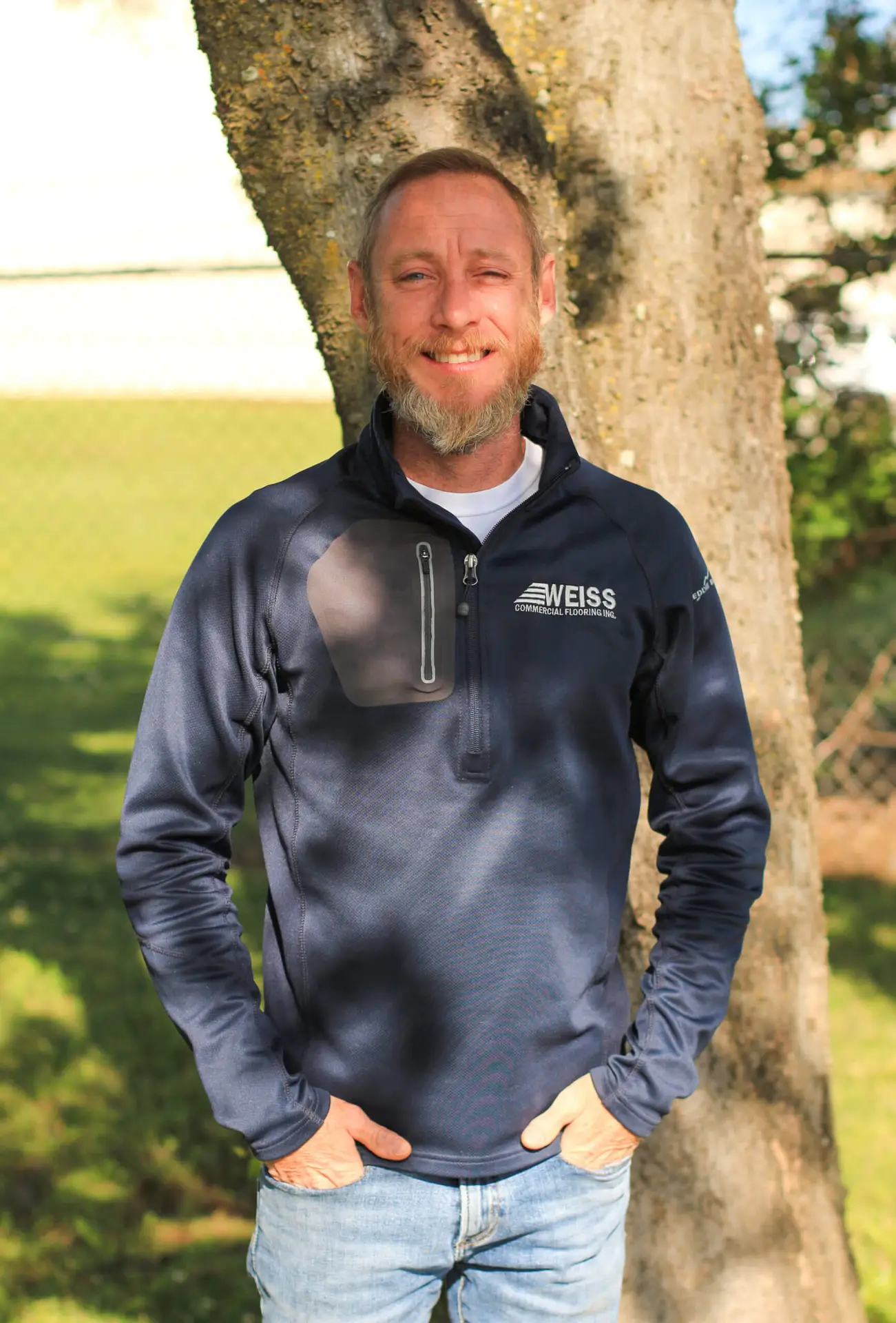 Adam Weiss owner of Weiss Commercial Flooring in Montgomery, AL standing in front of a tree, smiling, wearing a blue Weiss pullover